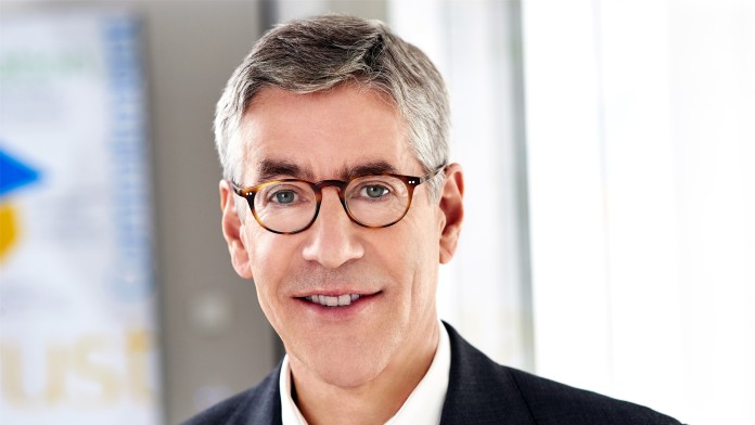 Porträt eines Mannes mit grauen Haaren, Brille und dunklem Sakko vor unscharfem Hintergrund