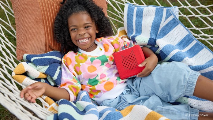 Ein kleines Mädchen liegt mit einer Toniesbox in seinen Armen auf Kissen.