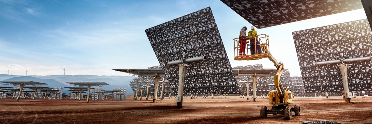 Zwei Personen auf einer Hebebühne vor großen, rechteckigen Solarpanelen mit Muster, die auf einem offenen Gelände aufgestellt sind