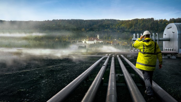 Person in gelber Schutzkleidung und Helm geht auf mehreren großen Rohren in einer nebligen Industrieanlage mit bewaldetem Hintergrund