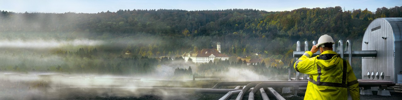 Person in gelber Schutzkleidung und Helm geht auf mehreren großen Rohren in einer nebligen Industrieanlage mit bewaldetem Hintergrund