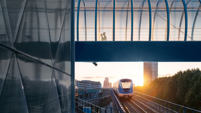 Zug auf Gleisen unter einer Fußgängerbrücke mit zwei Personen darauf und Sonnenuntergang im Hintergrund