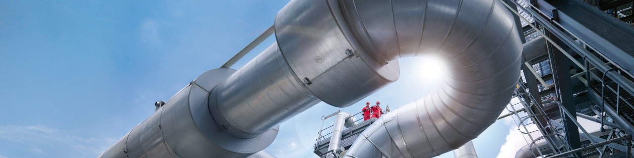 Große silberne Rohrleitungen in einem Industriekomplex vor blauem Himmel mit zwei Personen in roter Schutzkleidung auf einem Steg