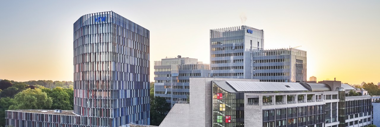 Drohnenaufnahme des KfW Gebäudes in Frankfurt bei Sonnenuntergang. Im Vordergrund eine Straßenkreuzung, links das IPEX Gebäude, rechtes die Nordarkade der KfW, im Hintergrund das Haupthaus der KfW.