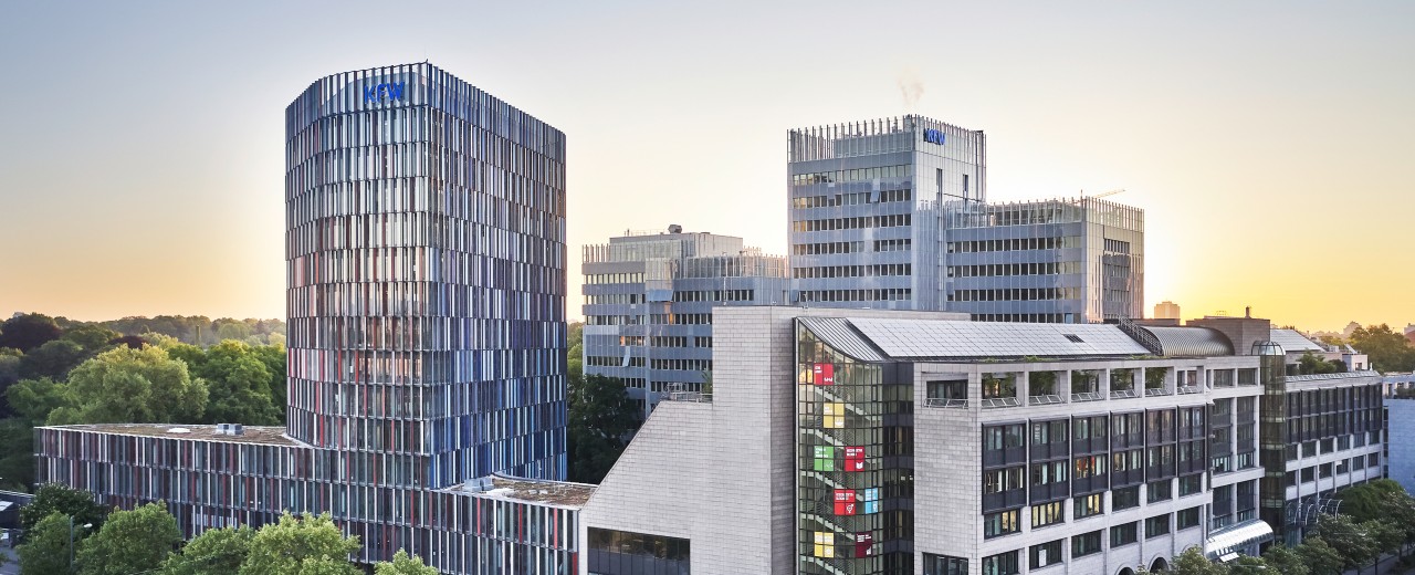 Drohnenaufnahme des KfW Gebäudes in Frankfurt bei Sonnenuntergang. Im Vordergrund eine Straßenkreuzung, links das IPEX Gebäude, rechtes die Nordarkade der KfW, im Hintergrund das Haupthaus der KfW.