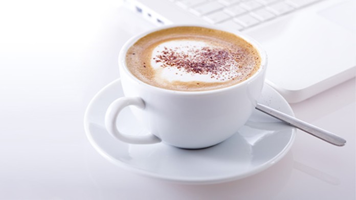 Eine Tasse mit Cappuccino steht vor einem Laptop. 
