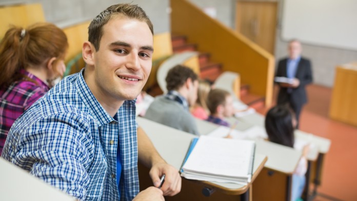 Studenten sitzen im Hörsaal. Ein Student schaut nach hinten und lächelt in die Kamera.
