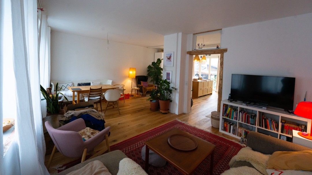 Blick in den großzügigen Wohnbereich eines sanierten Hauses, mit Sofa, Sessel, Fernseher auf einem Sideboard sowie einer Essecke