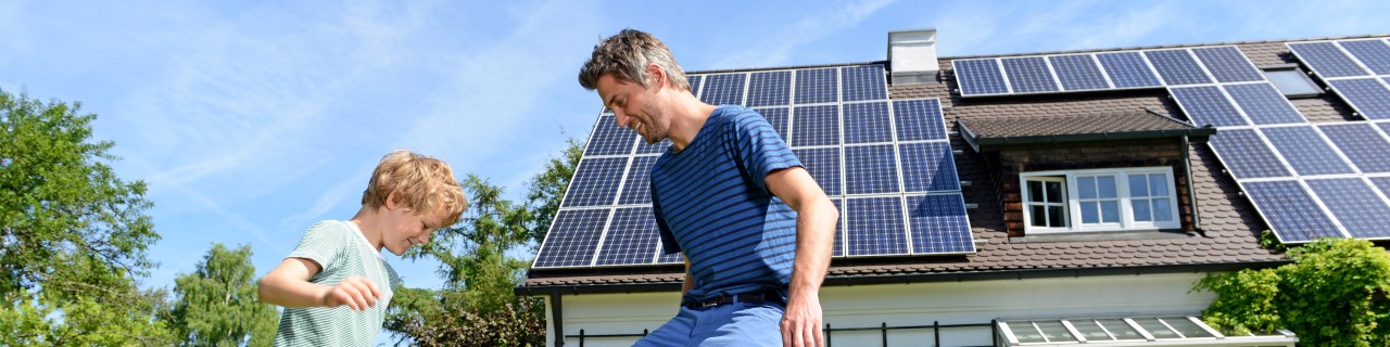 Bestehendes Einfamilienhaus mit Photovoltaikanlage auf dem Dach, Vater und Sohn im Vordergrund beim Fussball spielen.