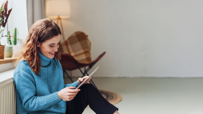 junge Frau sitzt entspannt auf dem Boden unter dem Fenster an der Heizung neben einem Sessel und schaut lächelnd auf ein Tablet-PC