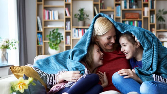 eine ältere Frau sitzt mit zwei Kindern fröhlich lachend auf einem Sofa im Wohnzimmer, soe haben eine Decke quer über ihren Schultern