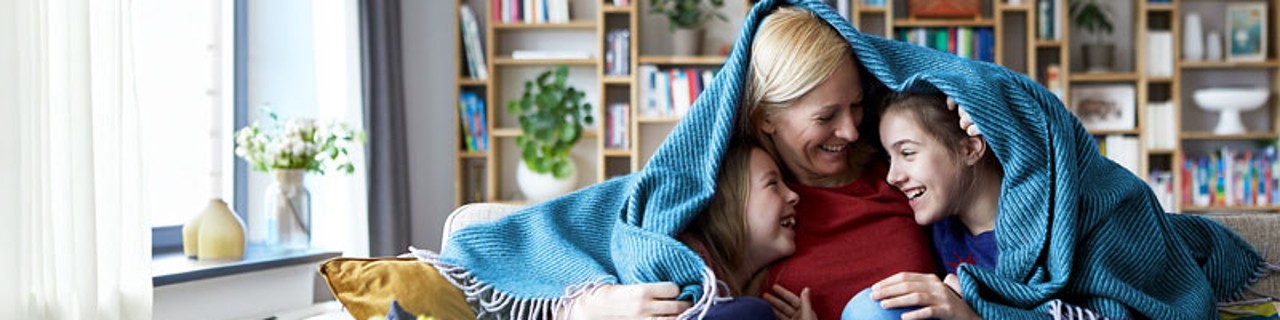 eine ältere Frau sitzt mit zwei Kindern fröhlich lachend auf einem Sofa im Wohnzimmer, soe haben eine Decke quer über ihren Schultern