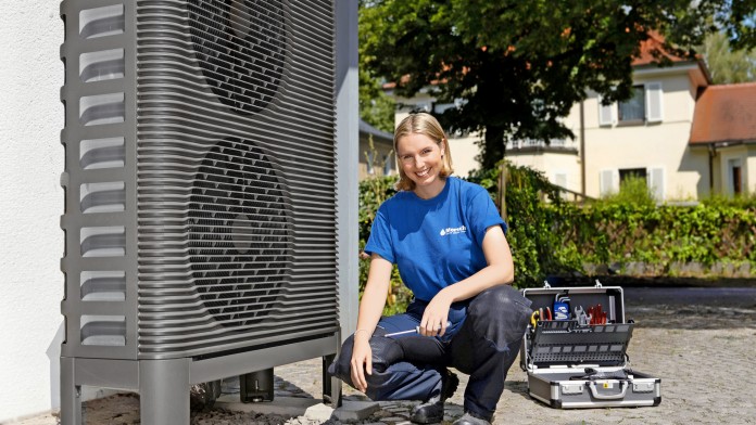 Die Anlagenmechanikerin kniet vor einer Wärmepumpe mit Werkzeug in der Hand.