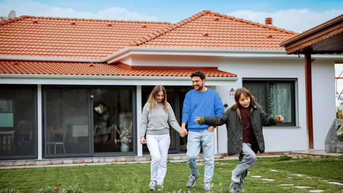Eine Familiesteht vor ihrem neu gekauftem Haus, einer Bestandsimmobilie und das Kind hüpft vergnügt vor den beiden Eltern über die Wiese des Vorgartens