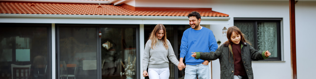 Eine Familiesteht vor ihrem neu gekauftem Haus, einer Bestandsimmobilie und das Kind hüpft vergnügt vor den beiden Eltern über die Wiese des Vorgartens