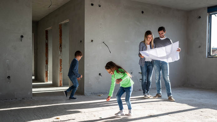 Eine Familie mit zwei Kindern steht in ihrem Neubau, die Elten halten die Ausbaupläne in den Händen und betrachten sie. Der kleine Junge und das Mädchen tollen im Raum herum, das Mädchen spielt in der Sonne, die durch die großen Fensteröffnungen fällt.