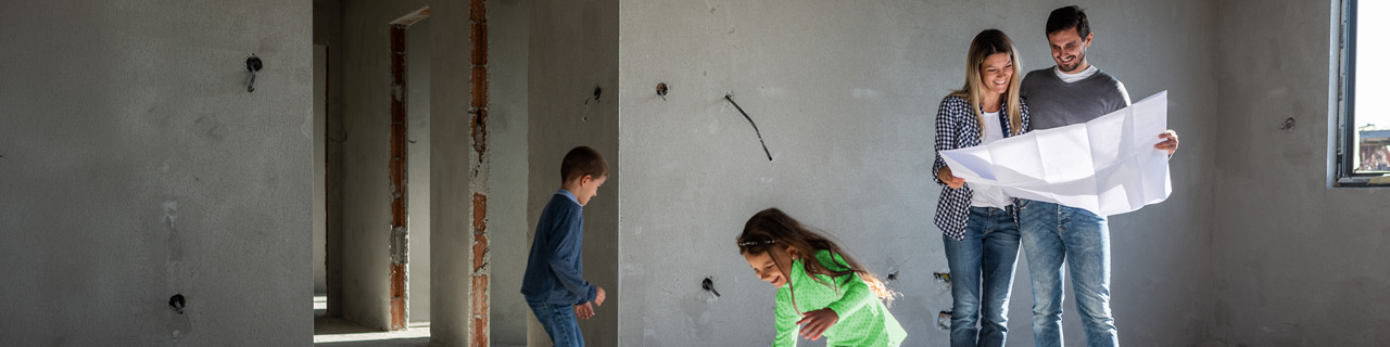 Eine Familie mit zwei Kindern steht in ihrem Neubau, die Elten halten die Ausbaupläne in den Händen und betrachten sie. Der kleine Junge und das Mädchen tollen im Raum herum, das Mädchen spielt in der Sonne, die durch die großen Fensteröffnungen fällt.