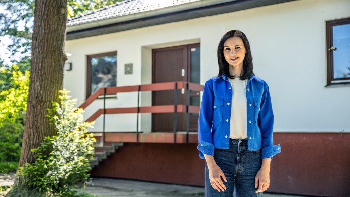 Junge Frau steht selbstbewusst vor einem Haus.