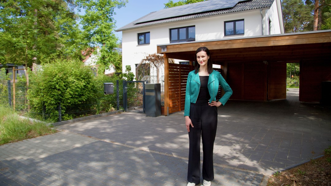 eine Frau steht entspannt vor einem Einfamlienhaus mit Solaranlage auf dem Dach und einem Carport