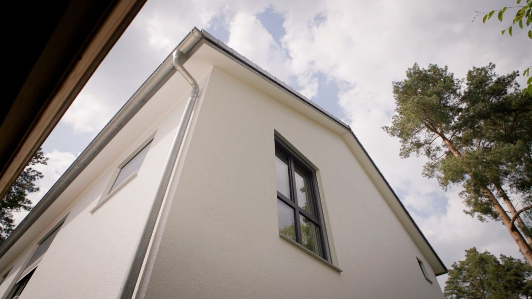 Aufsicht entlang einer weißen Hausfassade zum Schrägdach, neben dem Haus stehen einige Bäume