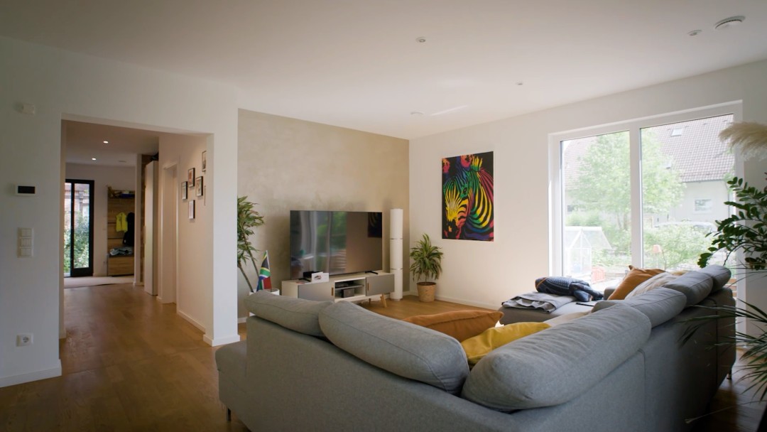 Blick in ein Wohnzimmer, über eine große Couch hinweg, mit Blick in einen Flur, auf einen Fernseher und aus dem Fenster