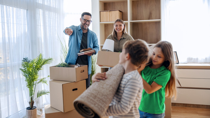 Eine Famile baut ihr neues Zuhause um bzw. räumt nach dem Umbau wieder ein - die beiden kleinen Kinder, ein Junge und ein Mädchen, tragen den Teppich über ihren Schultern, die Eltern zeigen an, wo er hinsoll. 