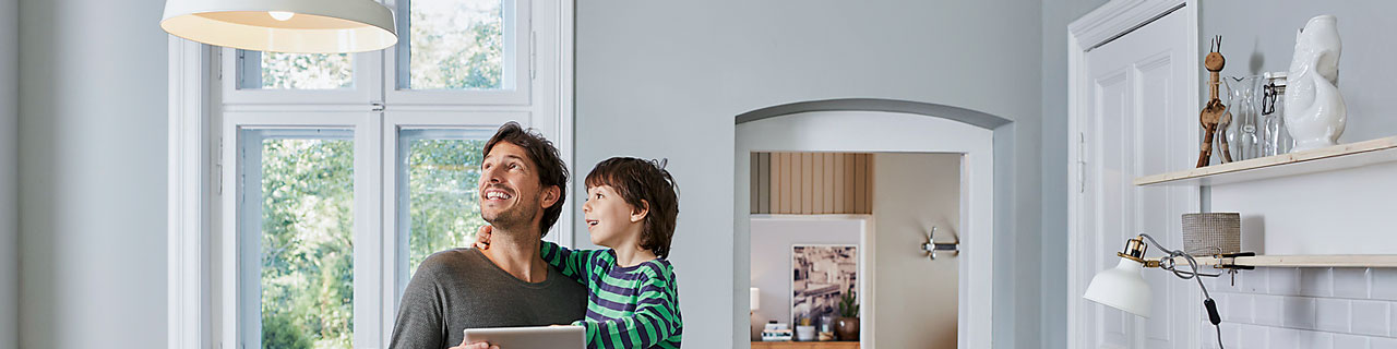 Vater mit Kind auf dem Arm, schauen beide begeistert die Lampe an, welche über das Tablett gesteuert wird.