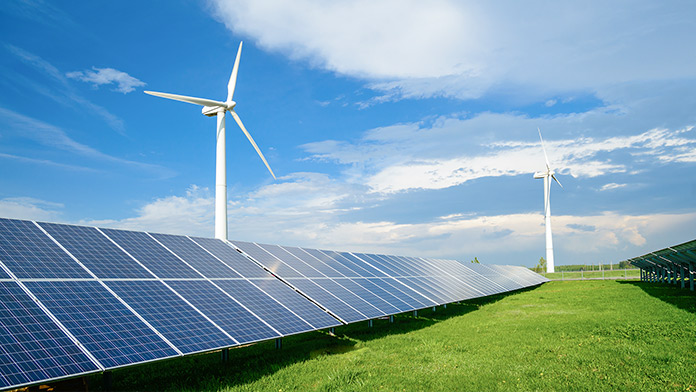 Solar panels on a green field, behind them 2 wind turbines under blue sky