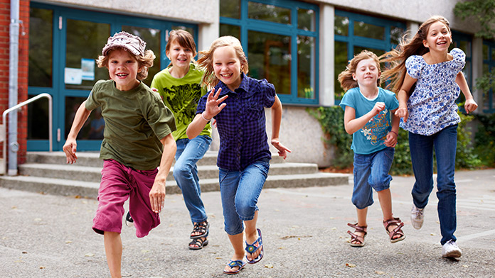 Vor einem Schulgebäude rennen 5 lachende Grundschulkinder der Kamera entgegen