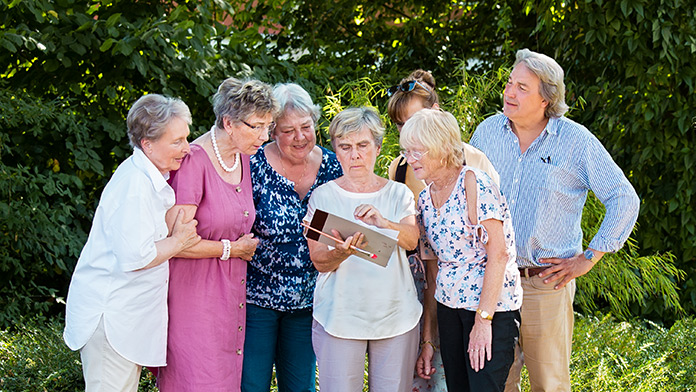 Gruppe stehender Senioren im Garten, die auf die Bilder von 2 Malerinnen blicken