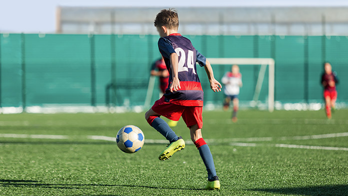 Fussballplatz vor einer Halle, wo ein Junge im Trikot den Ball zum Tor dribbelt