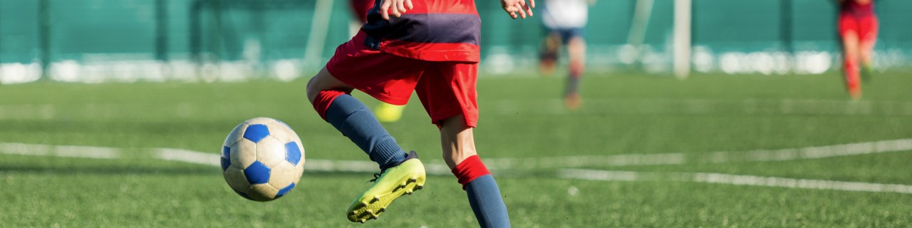 Fussballplatz vor einer Halle, wo ein Junge im Trikot den Ball zum Tor dribbelt