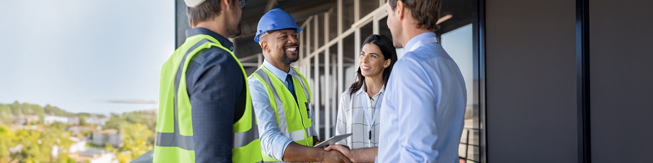 Zwei Energieeffizienz-Experten mit blauem Bauarbeiterhelm und gelber Weste sowie eine Frau und ein Mann in Business-Kleidung. Es werden Hände geschüttelt.