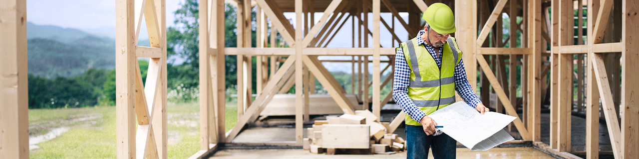 Ein Bauarbeiter steht mit Bauplänen in einem Rohbau aus Holz