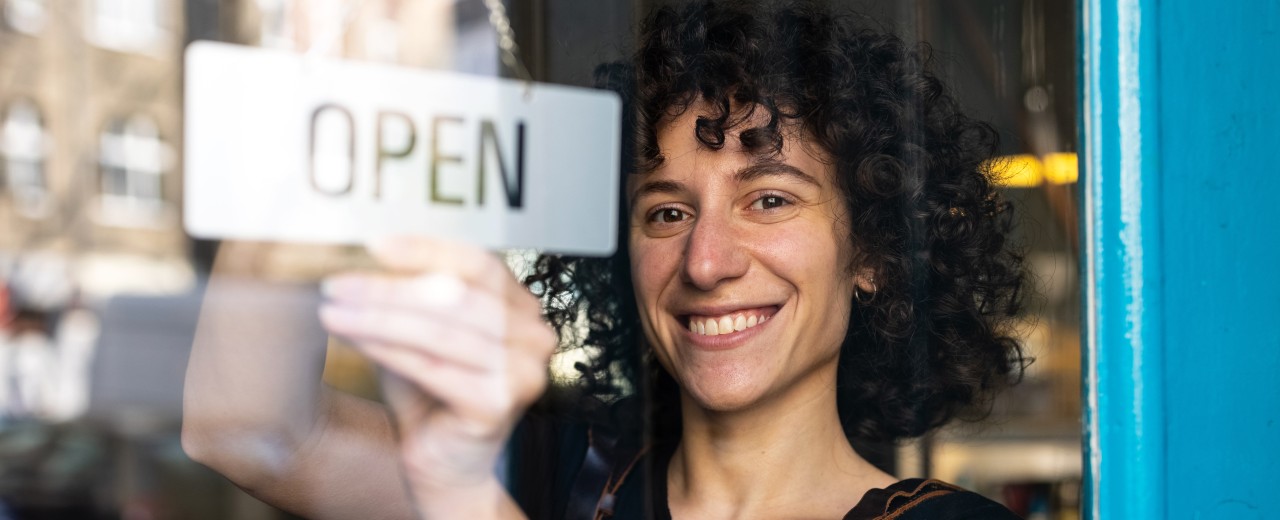 Eine lächelnde Frau mit Open-Schild an einer Ladentür