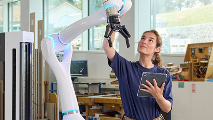 Eine junge Frau hält ein Tablet in der Hand und arbeitet an einem weißen Industrieroboterarm.