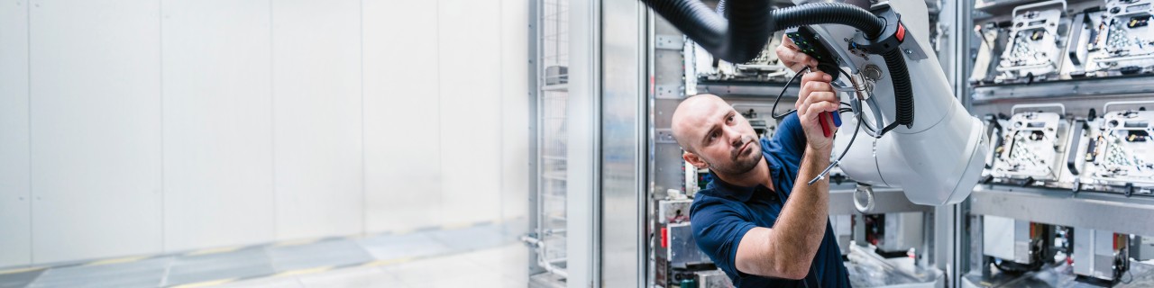 Mann mit blauem T-Shirt arbeitet an einem großen weißen Industrieroboterarm in einer hellen Werkhalle