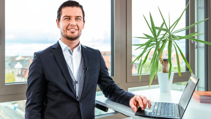 Ein lächelnder Mann mit Anzug steht in einem Büro an seinem Laptop