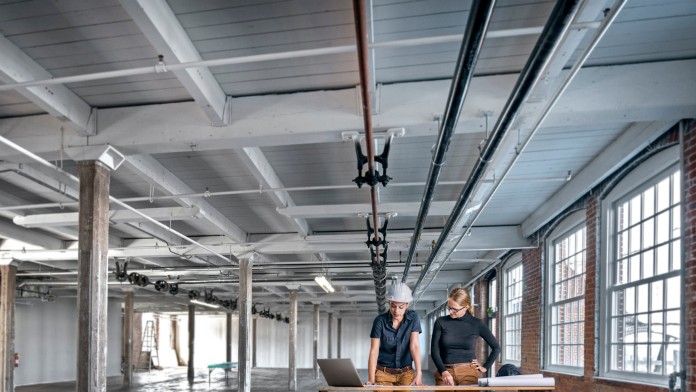 Zwei Frauen stehen mit Bauplänen und Laptop in einem industriellen Gebäude