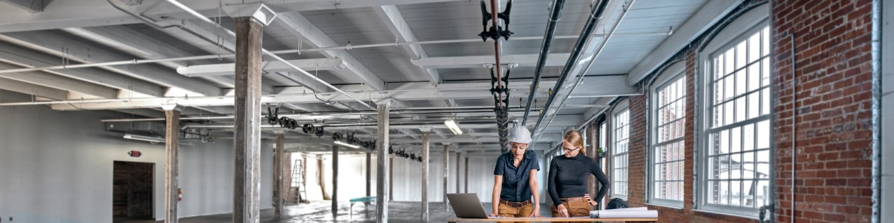 Zwei Frauen stehen mit Bauplänen und Laptop in einem industriellen Gebäude