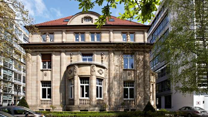 Außenansicht der Villa 102, ein neoklassizistisches Gebäude. Im Vordergrund ist ein grüner Rasen. Links und rechts sind Bürogebäude.