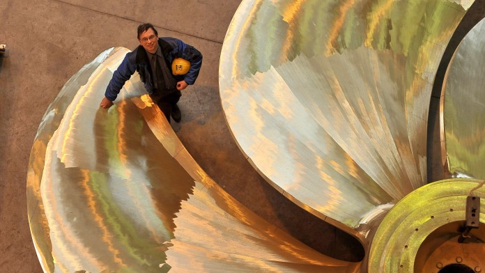 Managing director of a ship propeller manufacturer stands next to a large propeller in a workshop