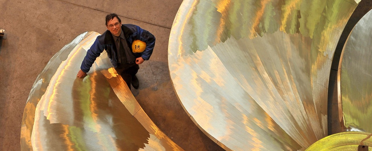 Managing director of a ship propeller manufacturer stands next to a large propeller in a workshop