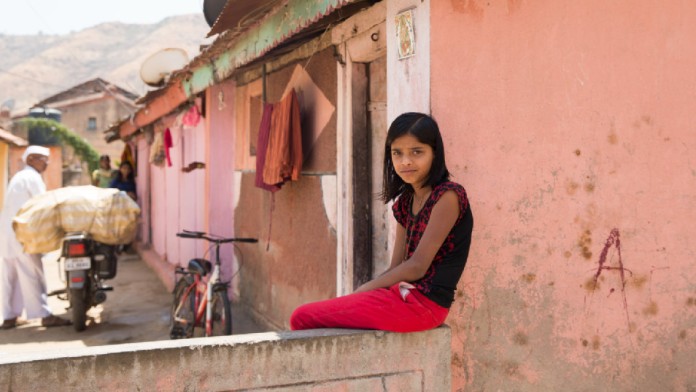 An Indian girl sits on a wall by a house on a village street and looks smilingly into the camera. In the background, a man pushes a motorbike loaded with goods.