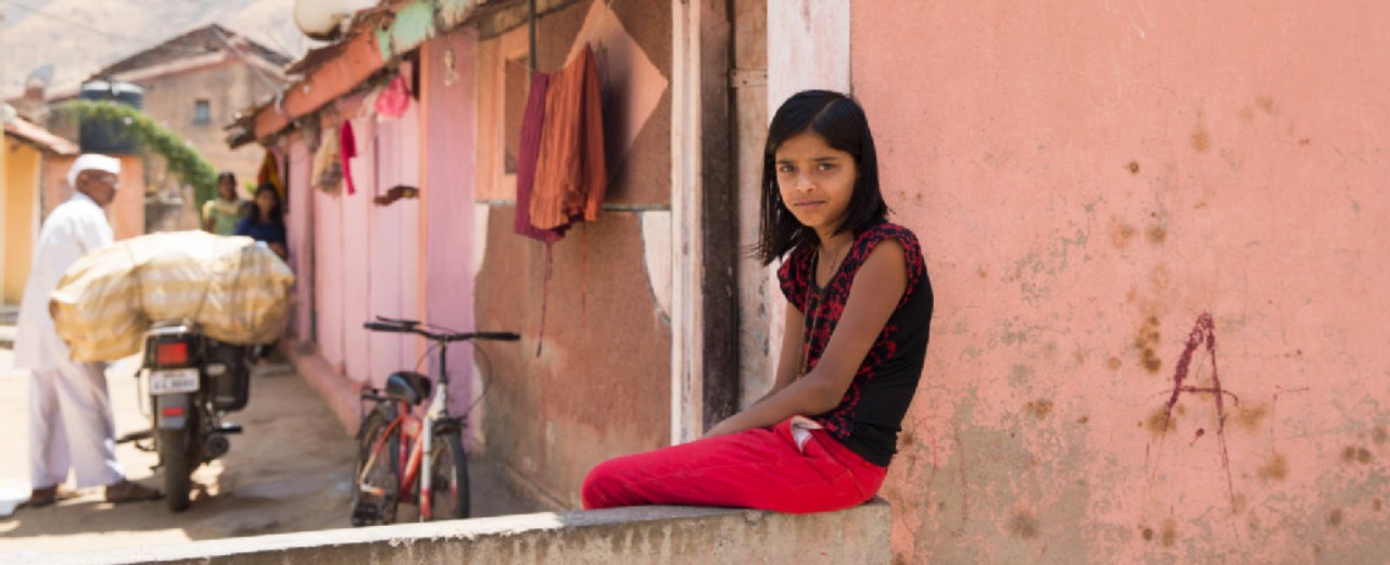 An Indian girl sits on a wall by a house on a village street and looks into the camera with a smile. In the background, a man pushes a bicycle.