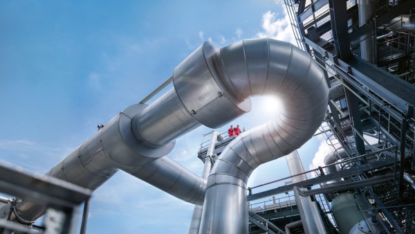 Large silver pipes in an industrial complex against a blue sky, with two people in red protective clothing on a walkway