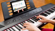 Hände auf E-Piano-Tastatur, auf dem Notenhalter steht ein iPad, auf dem Display sieht man eine Skoove-Lektion