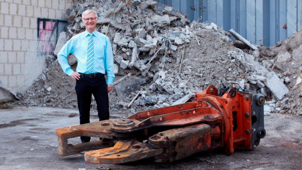 Illingens Bürgermeister Armin König posiert neben einer Abrisszange vor einem Schutthaufen