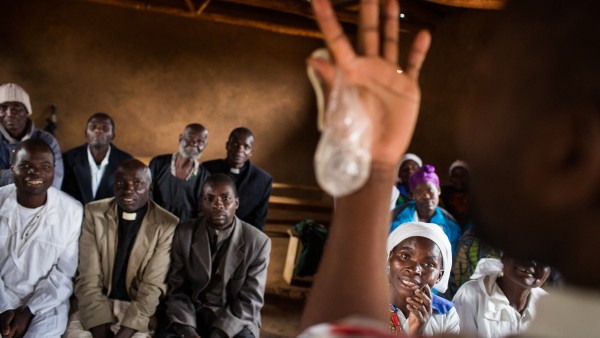 Ein Kondom wird vor einer Gruppe von Priestern und anderen Zuhörern hochgehalten