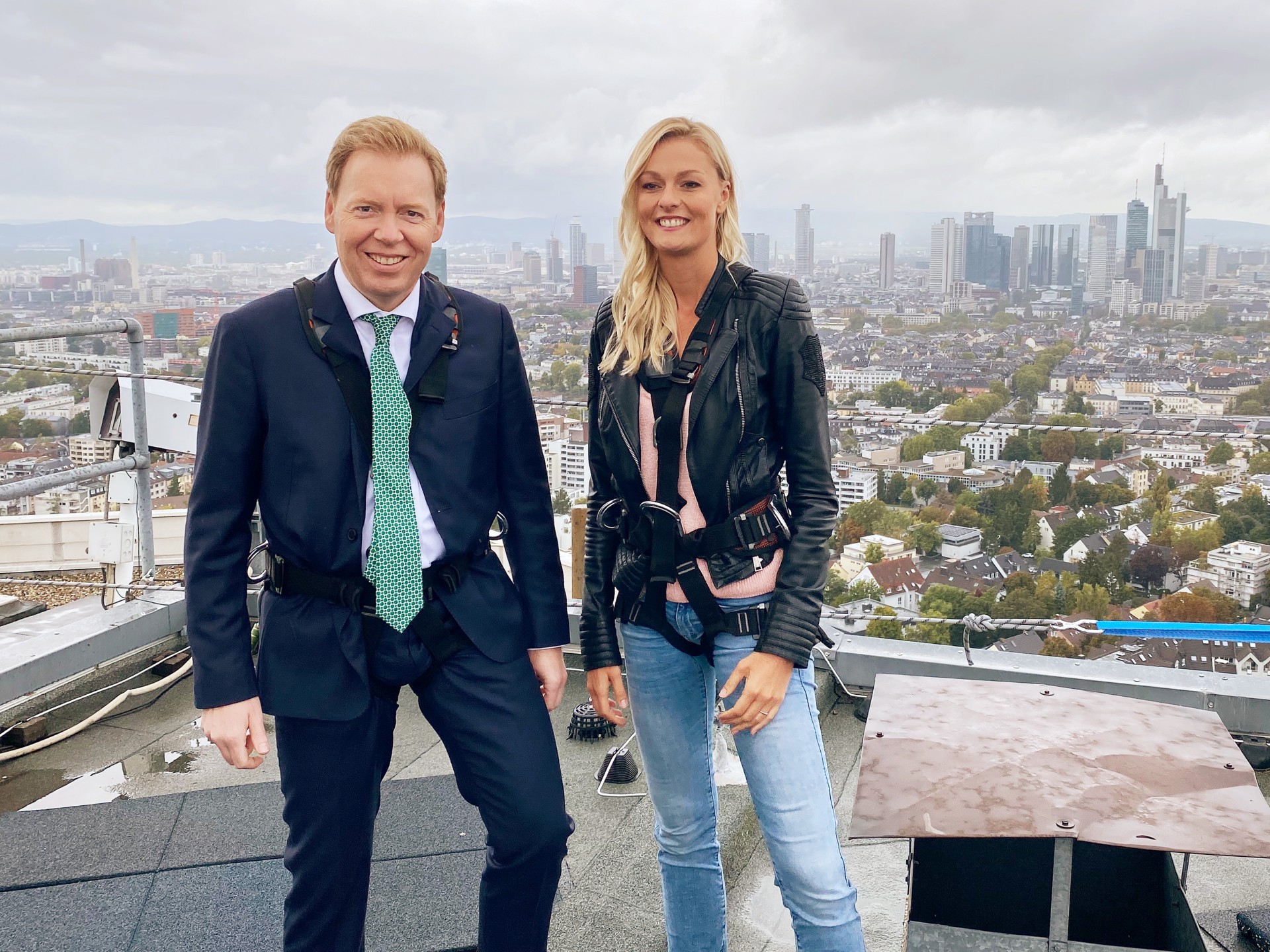 Man in suit and woman in casual clothes stand smiling on a skyscraper roof